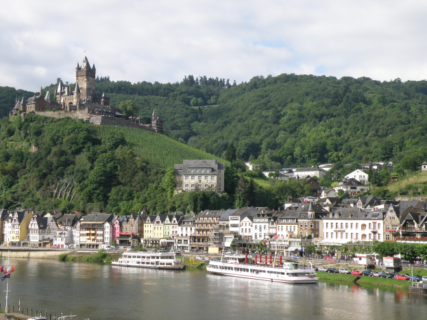 Ein malerischer Blick auf den Rhein in Deutschland, mit einer Burg auf einem Hügel im Hintergrund, Booten auf dem Fluss, Fahrzeugen auf einer nahen Straße und einem bewölkten Himmel.