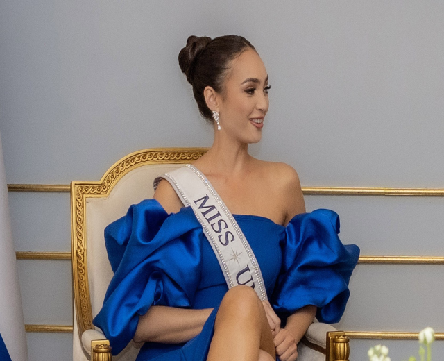 Frau in einem blauen Kleid mit einer "Miss Universe 2019" Schärpe, die auf einem Stuhl sitzt und von Blumen umgeben ist.