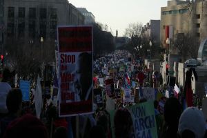 Menschen protestieren auf einer Straße und halten Plakate, mit Gebäuden, Bäumen, Laternenmasten und dem Himmel im Hintergrund.