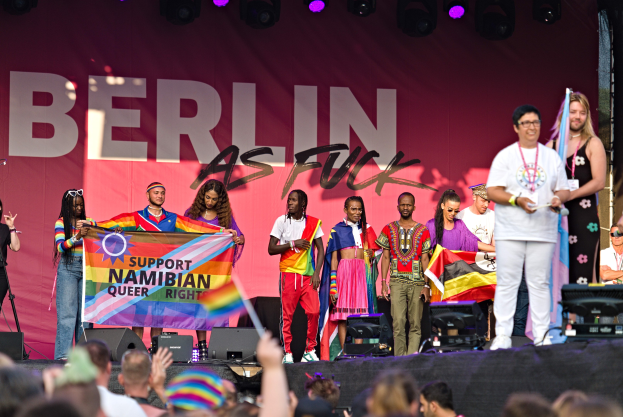 Gruppe von Menschen auf einer Bühne mit einem Banner, auf dem 'Berlin as Fuck' steht, mit Lautsprechern und einer Menge unten, die Hände hebt.
