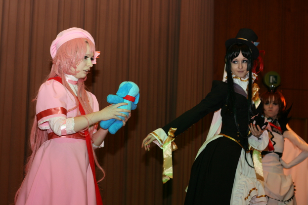 Drei Frauen sind in einer dramatischen Performance zu sehen, wobei eine in einem pinkfarbenen Kleid mit einem blauen Teddybär und eine andere in einem schwarzen Kleid, die nach links schaut, sowie ein brauner Vorhang im Hintergrund.