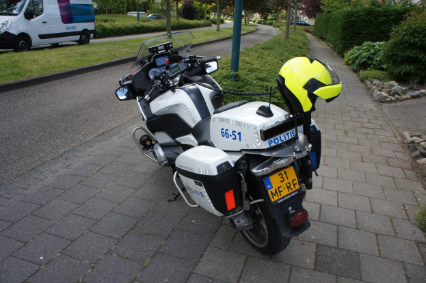 Ein Polizeimotorrad am Straßenrand mit einem Helm darauf, umgeben von Bäumen, Pfählen, Gebäuden, Pflanzen, Gras, Steinen und anderen Fahrzeugen.