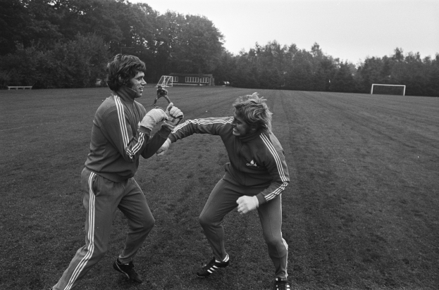 Zwei Männer in Schwarz-Weiß-Uniformen sind in eine körperliche Auseinandersetzung auf einem Fußballfeld verwickelt, wobei Bäume und ein Schuppen im Hintergrund zu sehen sind.