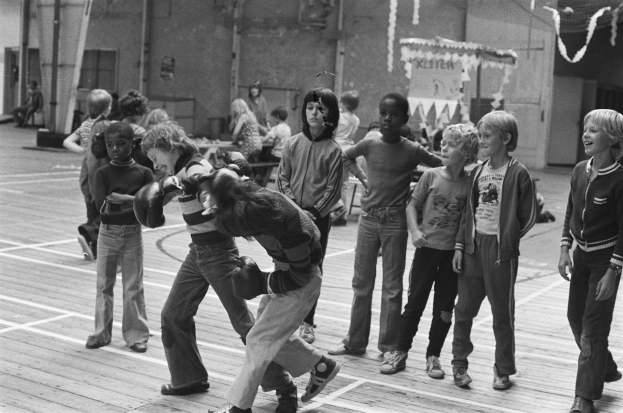 Eine Gruppe von Kindern, die auf einem Basketballfeld spielen, mit zwei kämpfenden Personen in der Mitte und anderen auf Bänken im Hintergrund, dargestellt in Schwarz-Weiß.