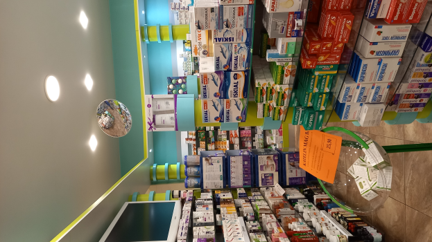 Eine Apotheken-Theke mit verschiedenen Medikamenten in Schachteln und Flaschen auf Gestellen, einem Spiegel an der Wand, Deckenlampen und einem Glastisch mit Papieren und Gegenständen darauf, der Boden ist unten sichtbar.