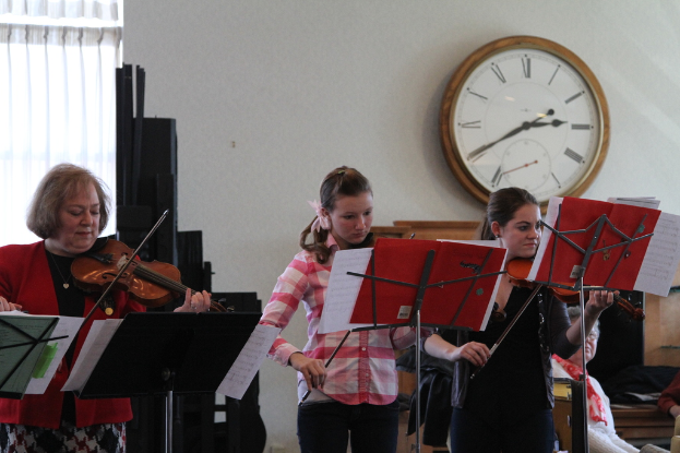 Drei Frauen auf der B├╝hne stehend und Geigen spielend, mit Notenpulten und Notenbl├Ąttern davor, w├Ąhrend ein Zuhörer sitzt und zuh├Ârt, mit einer Uhr an der Wand und einem wei├čen Vorhang im Hintergrund.