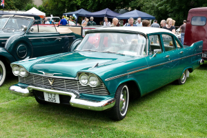 Alte Autos auf Gras geparkt mit Menschen dahinter, Zelte im Hintergrund, Bäume oben und eine Flagge oben links.