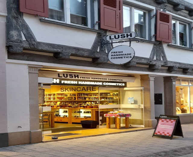 Außenansicht eines "Lush Skincare"-Ladens mit sichtbarer Innenausstattung, Schreibtisch, Stuhl und Bodenbelag.