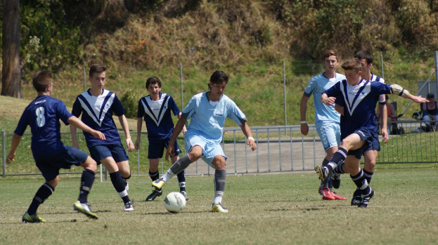 Eine Gruppe von Menschen spielt Fußball mit einem Ball auf dem Boden, während zwei Personen im Hintergrund auf Stühlen sitzen und weiter weg Bäume zu sehen sind.