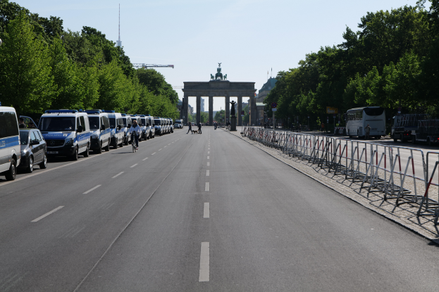 Eine lange Reihe von Polizeiwagen steht am Straßenrand vor dem Brandenburger Tor in Berlin, Deutschland, mit Menschen, die Fahrräder fahren und auf der Straße stehen, Barrieren, Bäumen und einem Bogen mit Statuen im Hintergrund.