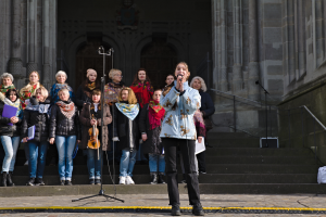 Eine Gruppe von Menschen, einige mit Musikinstrumenten, steht vor einem Gebäude mit Treppen und Geländern, während eine Frau in der Mitte in ein Mikrofon singt, umgeben von zusätzlichen Mikrofonen auf Ständern.