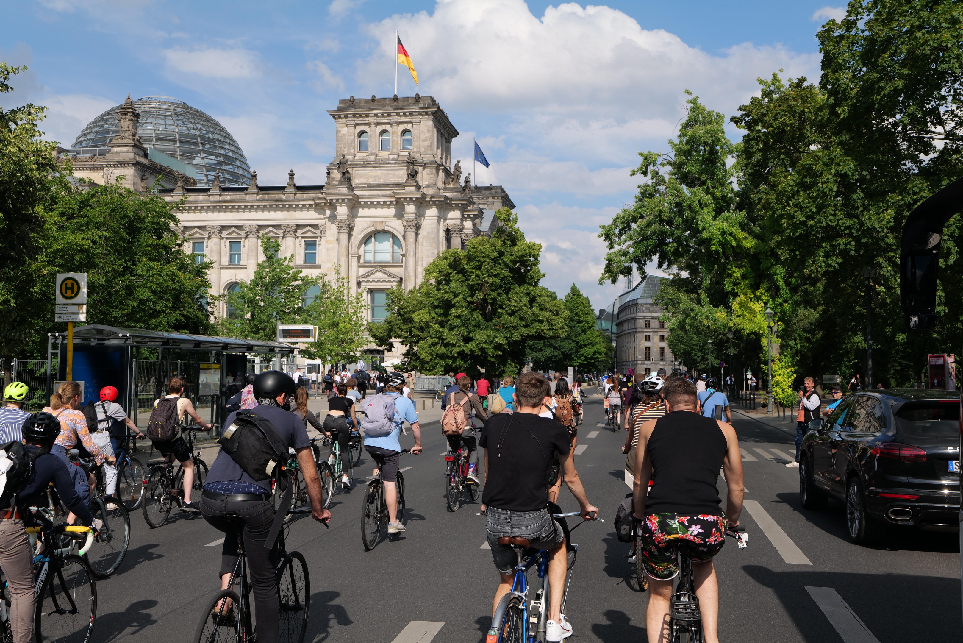 Eine Gruppe von Menschen, die auf Fahrrädern eine von Bäumen gesäumte Straße in Berlin entlangfahren, mit Gebäuden und einer Bushaltestelle im Bild, unter einem bewölkten Himmel und einer Flagge auf einem der Gebäude.
