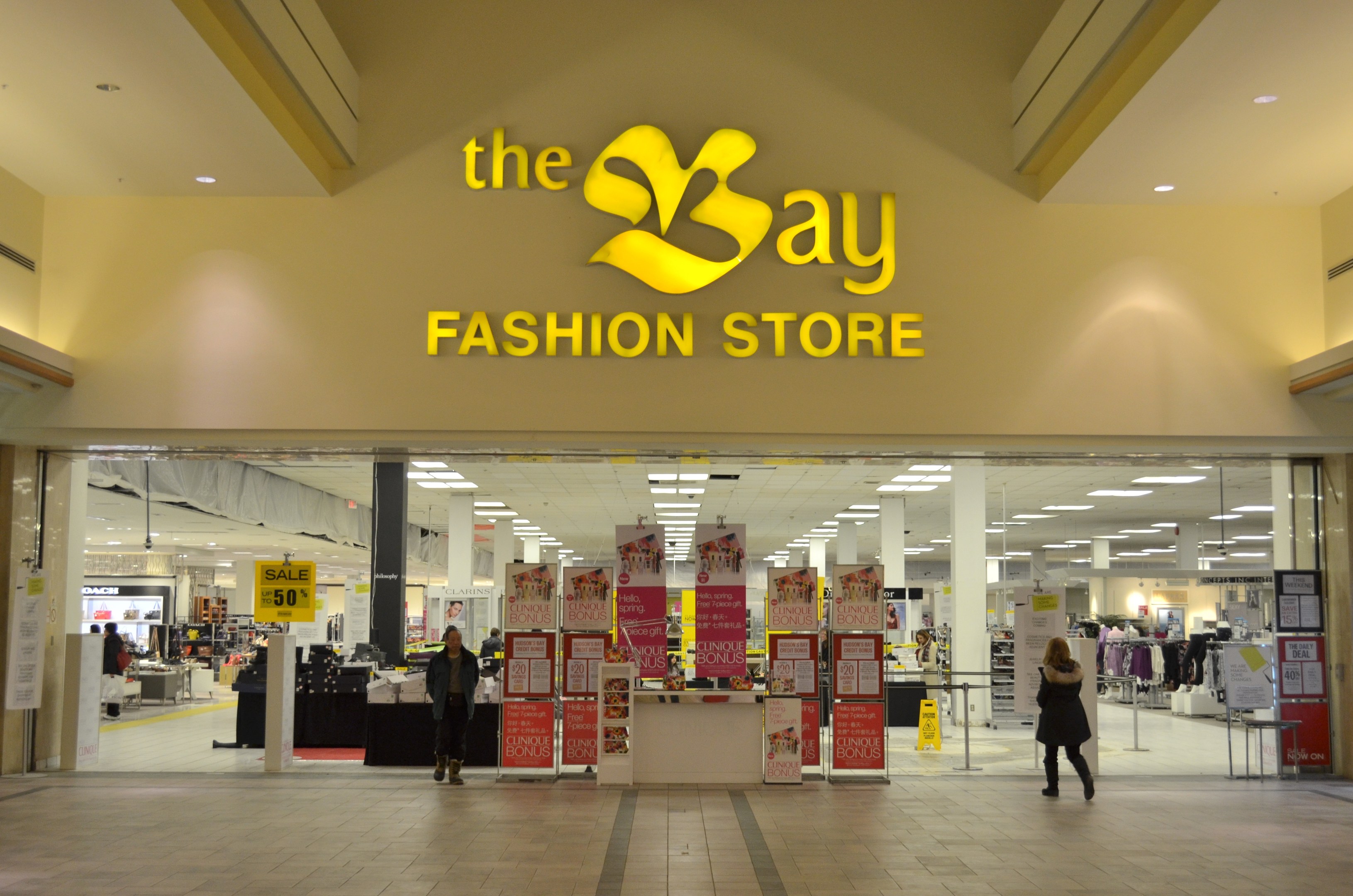 Außenansicht eines Bay Fashion-Geschäfts in einem Einkaufszentrum mit Einkäufern, Wandtafeln, Säulen, Deckenleuchten und Hintergrundauslagen.