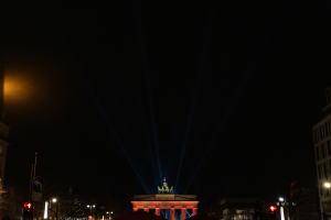 Brandenburger Tor in Berlin bei Nacht, beleuchtet von Straßenlaternen, mit umliegenden Gebäuden, Bäumen, Pfählen, Verkehrszeichen, Schildern und Zelten gegen einen dunklen Himmel.
