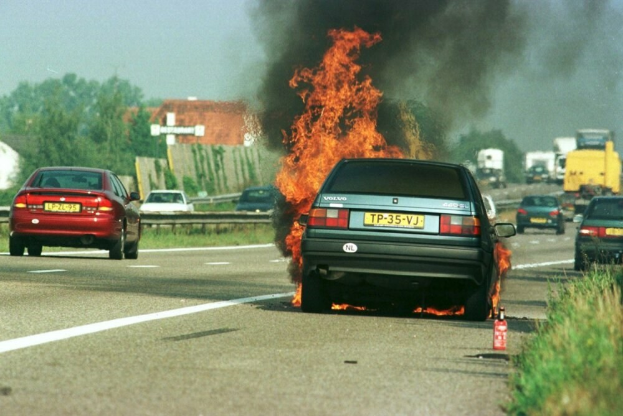 Ein Auto in Flammen auf dem Seitenstreifen, umgeben von anderen Fahrzeugen, mit B├Ąumen, Geb├Ąuden und einem klaren blauen Himmel im Hintergrund und Gras und einem Feuerl├Âscher auf der rechten Seite.