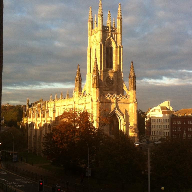 Eine Straße mit Verkehr und Ampeln, eine Person, ein Metallzaun und im Hintergrund Bäume, Gebäude und Himmel.