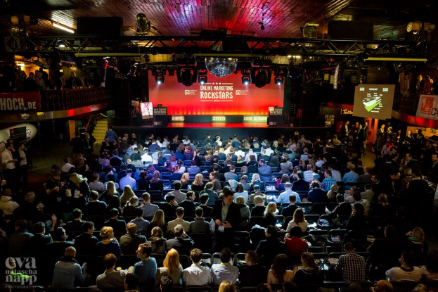 Eine große Menschenmenge sitzt auf Stühlen vor einer Bühne mit Bildschirmen und Plakaten, die von Scheinwerfern beleuchtet und von Geländern umgeben sind und gespannt auf den Beginn eines Online-Marketing-Rockstars-Events warten, mit einem Wasserzeichen in der linken unteren Ecke.