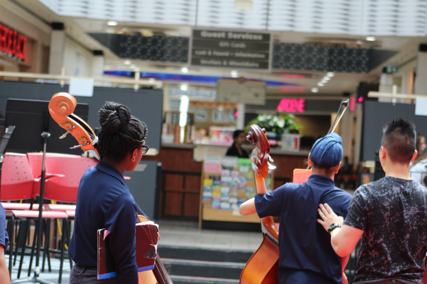 Eine Gruppe von Menschen, die Celli in einem Einkaufszentrum spielen, mit Stühlen, Notenpulten, Schildern und Lichtern im Hintergrund.