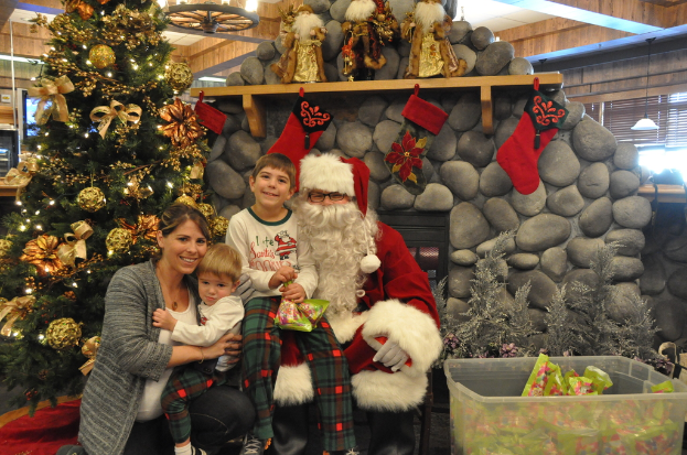 Eine Frau, Kinder und ein Mann in Santa-Klaus-Kostüm mit Brille und Handschuhen stehen neben einem geschmückten Weihnachtsbaum, Geschenken und einem Korb mit Gegenständen, mit einer Felswand und Lichtern im Hintergrund.