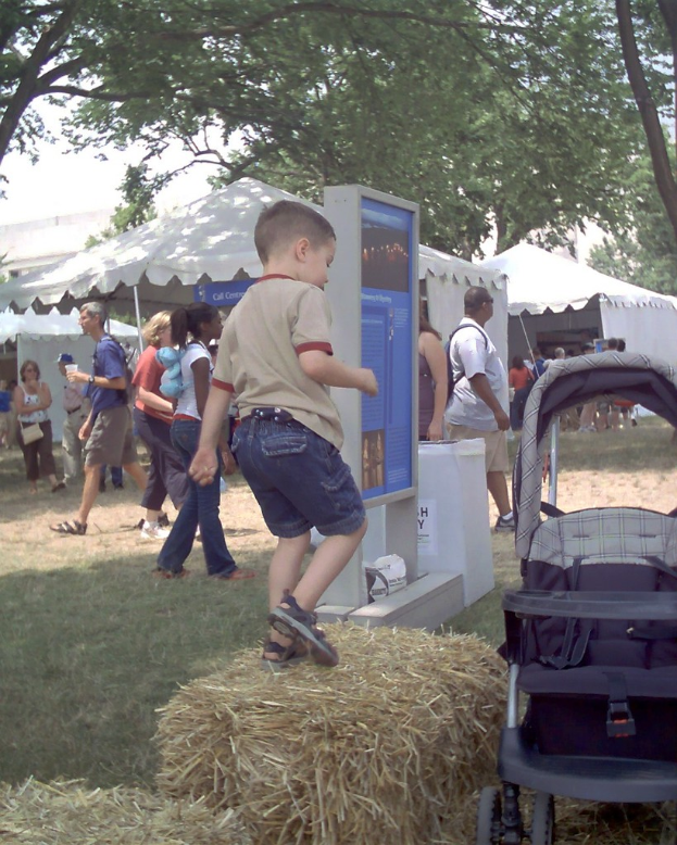 Eine Szene mit mehreren Zelten und Menschen, die darum herum gehen, ein Anzeigebrett vorne, trockenes Gras mit einem Kind darauf und Bäume auf beiden Seiten.