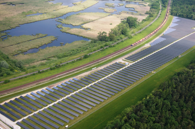 Luftaufnahme einer Solaränlage mit Panels, umgeben von Bäumen, Gras und Wasser, mit einem Zug auf einer näheren Eisenbahnschiene.