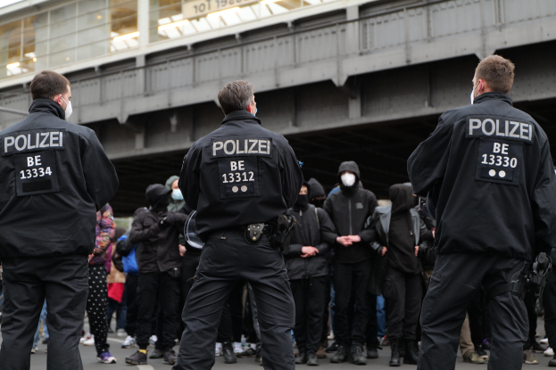 Eine Gruppe von Polizeibeamten in Uniform steht vor einer Menge maskierter Individuals in Schwarz, mit einer Stadtbrücke und einem Gebäude im Hintergrund, während einer Demonstration.