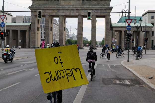 Eine Gruppe von Radfahrern mit Helmen fährt vor dem Brandenburger Tor in Berlin, Deutschland, vorbei, während einer eine gelbe Tafel hält, Lichtmasten, Verkehrszeichen, Gebäude, Bäume und einen klaren blauen Himmel im Hintergrund.