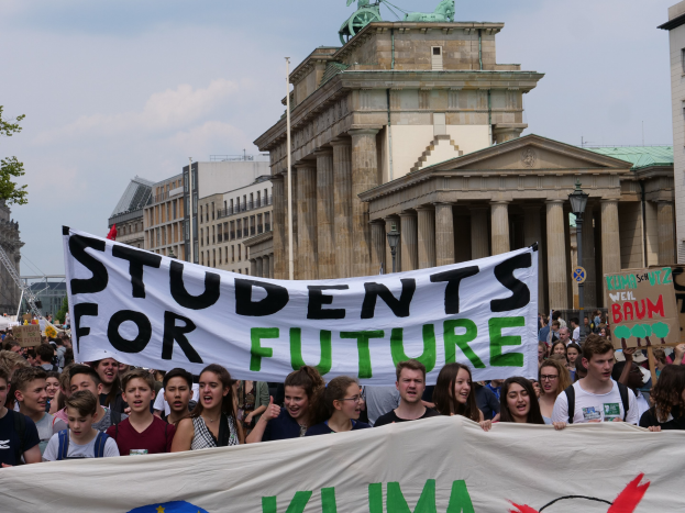 Gruppe von Schülern marschiert in Berlin mit einem bunt bemalten "Students for Future"-Schild gegen eine Kulisse aus Gebäuden, Bäumen und Himmel.