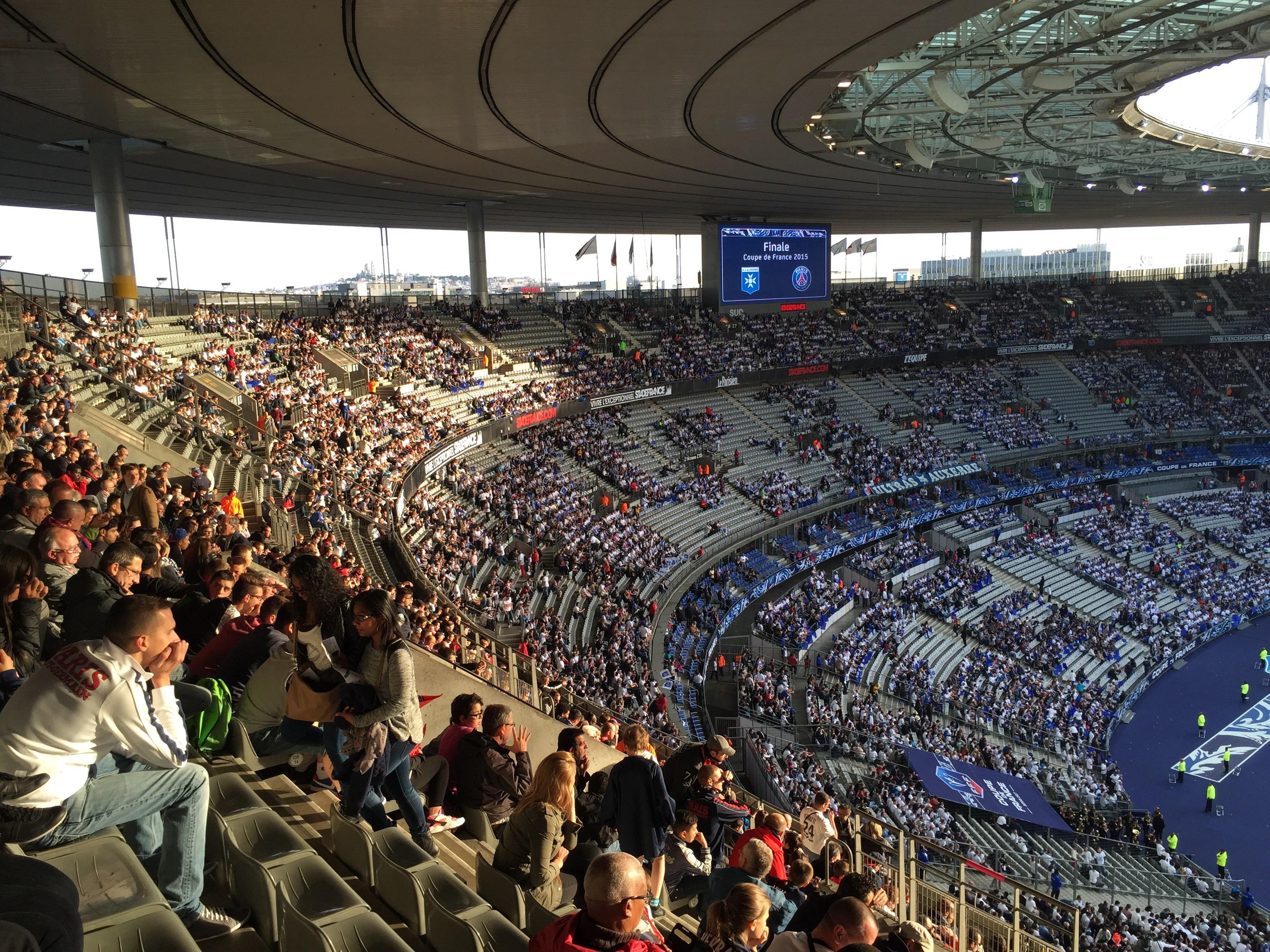 Große Menge in einem Stadion bei einem Fuballspiel mit einer Bühne, Fahnen, Masten und einem Bildschirm im Hintergrund, identifiziert als Allianz Arena in München, Deutschland.