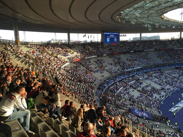 Große Menge in einem Stadion bei einem Fuballspiel mit einer Bühne, Fahnen, Masten und einem Bildschirm im Hintergrund, identifiziert als Allianz Arena in München, Deutschland.