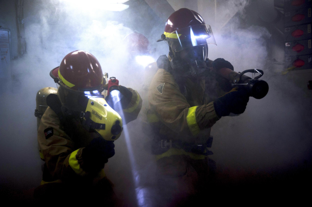 Zwei Feuerwehrleute in Schutzausrüstung und Gasmaske sprühen Wasser auf eine Wand mit Rauch, mit verschiedenen Gegenständen rechts und einem Textschild links.