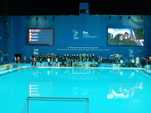 Gruppe von Menschen, die um ein Hallenbad mit Netz am Boden stehen, mit einer Wand im Hintergrund, die den Text und Logos "FINA World Championships 2015 Kazan" anzeigt, sowie Trägern und Lampen.