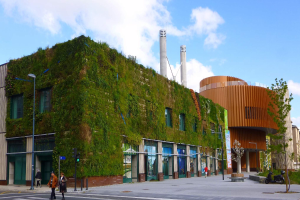 Ein großes Gebäude mit einer grünen Wand an der Seite, umgeben von Straßenmasten, Straßenlaternen, Verkehrspfosten, Ampeln, Kraftfahrzeugen, Fußgängern, Bäumen und einem bewölkten Himmel, als Teil des Green Building Projects.