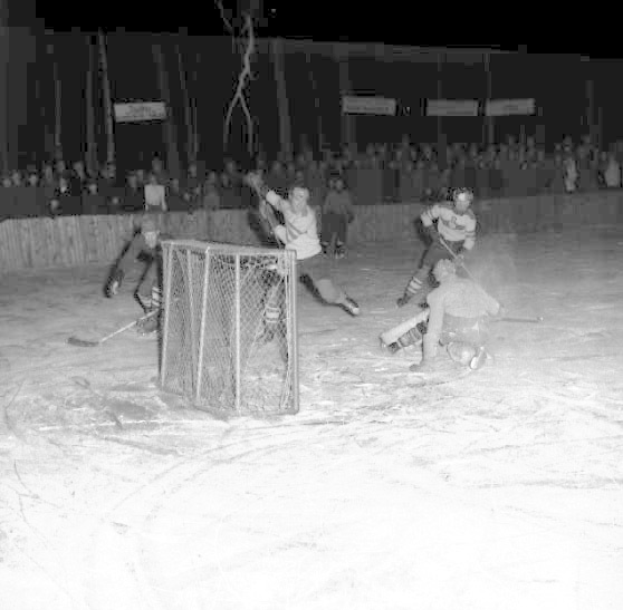 Schwarzes und weißes Foto von Menschen, die Hockey auf einem Eisplatz mit einem Netz im Vordergrund und Zuschauern im Hintergrund spielen.