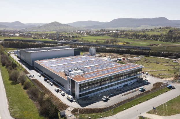 Luftaufnahme eines großen Gebäudes mit Solarpaneelen auf dem Dach, umgeben von Fahrzeugen, Bäumen, Gras, Masten und Hügeln unter einem klaren blauen Himmel, identifiziert als Deutschlands erste solarbetriebene Fabrik.