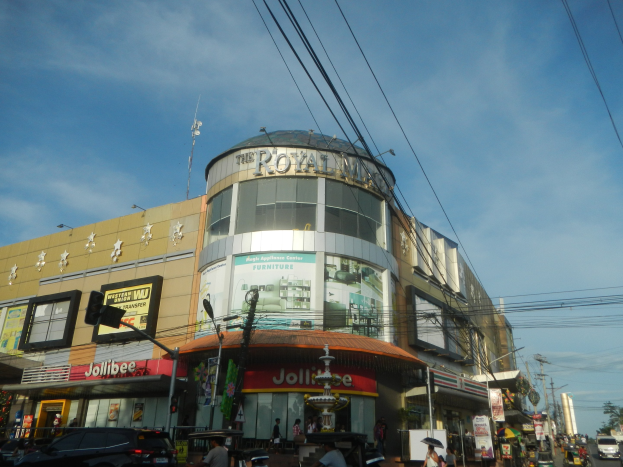 Eine belebte Stadtstraße mit fahrenden Fahrzeugen, Fußgängern auf dem Gehweg und dem Royal Mall Einkaufszentrum im Hintergrund, umgeben von Bäumen, Strommasten und Drähten unter dem Himmel.