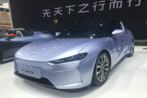 Ein silberner Peugeot K20 Wagen auf einer Automesse, umgeben von Menschen, mit einer schwarzen Texttafel im Hintergrund und Pflanzen auf der rechten Seite.