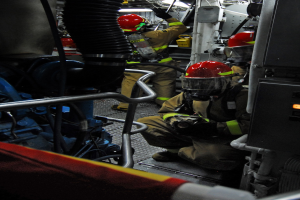 Feuerwehrleute in Helmen und Schutzausrüstung bei der Arbeit an einem Feuerwehrauto mit Rohren und einer Leuchte im Hintergrund.