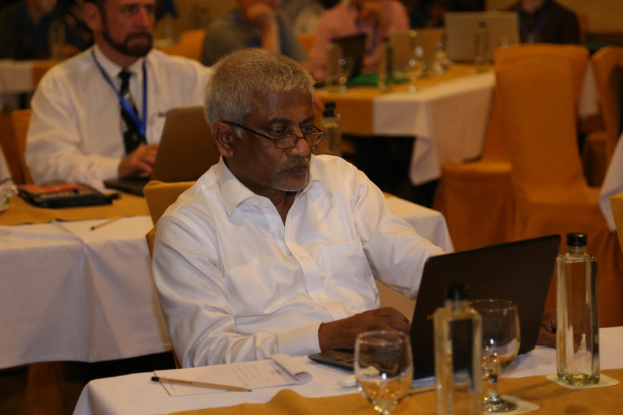 Ein Mann sitzt an einem Tisch und schaut auf einen Laptop, mit einem Weinglas und Flaschen in der Nähe, während ein anderer Mann im Hintergrund steht.