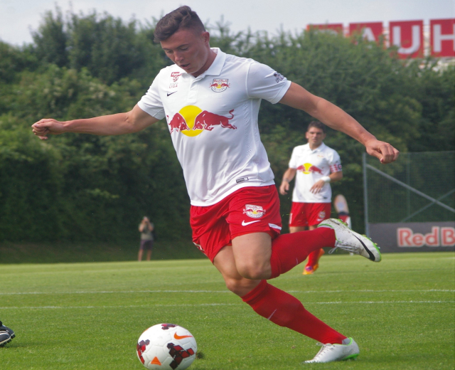 Ein professioneller Fußballspieler schießt einen Ball auf einem grünen Feld mit Bäumen und einem klaren blauen Himmel im Hintergrund, wobei "RB Leipzig" auf einer Tafel hinter ihm zu sehen ist.