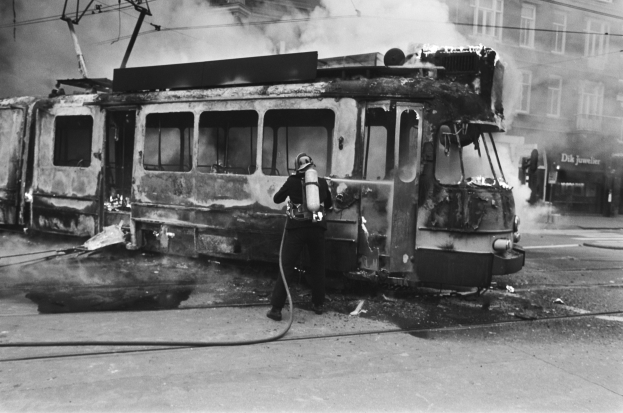 Schwarzes und wei├čes Bild eines Mannes in einem Helm, der eine Pfeife h├Ąlt und neben einer ausgebrannten Stra├čenbahn mit Rauch steht, vor dem Hintergrund eines Geb├Ąudes mit Fenstern und Dr├Ąhten.