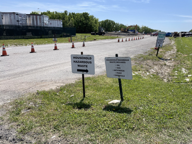 Ein Straßenschild mit der Aufschrift "Haushaltsgefährlicher Abfall" ist von Gras, Verkehrskegeln, Fahrzeugen, Bäumen und einem bewölkten Himmel umgeben.
