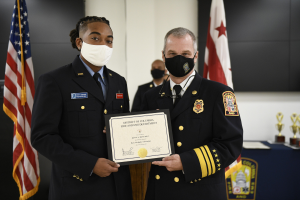 Zwei Männer in Feuerwachenuniformen stehen nebeneinander und tragen Masken und halten ein Zertifikat, mit einem Awards-Tisch und Flaggen im Hintergrund.