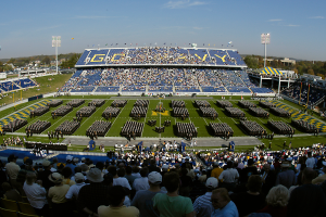 Eine große Menschenmenge in einem Stadion bei einem Fußballspiel, wobei einige stehen und andere sitzen, auf einem grasbewachsenen Feld unter einem klaren blauen Himmel mit Laternenmasten, Bäumen und Gebäuden im Hintergrund.