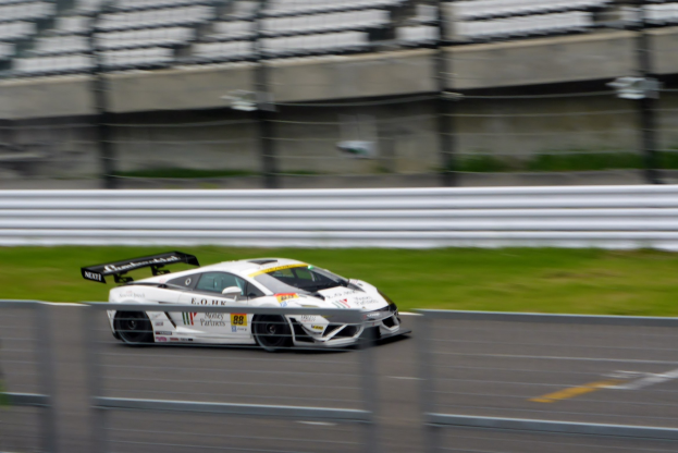Weißer Sportwagen fährt mit hoher Geschwindigkeit auf einer Rennstrecke, die von einem Zaun und Gras umgeben ist, mit einem unscharfen Hintergrund.