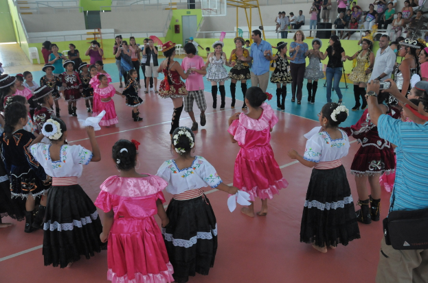 Eine Gruppe von Kindern in Kostümen, mit einem Mädchen und einem Jungen, die in der Mitte tanzen, während andere Menschen von der oberen rechten Seite des Bildes zuschauen.