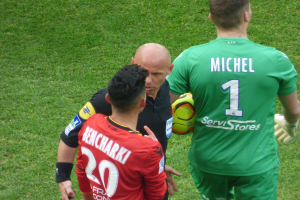 Schiedsrichter spricht mit zwei Fußballspielern auf einem saftig grünen Feld, einer hält den Ball.