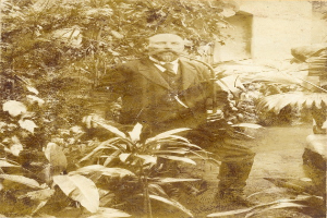 Altes Schwarz-Weiß-Foto eines Mannes in Anzug und Krawatte, der in einem Garten mit Pflanzen, Bäumen und einer Wand im Hintergrund steht.