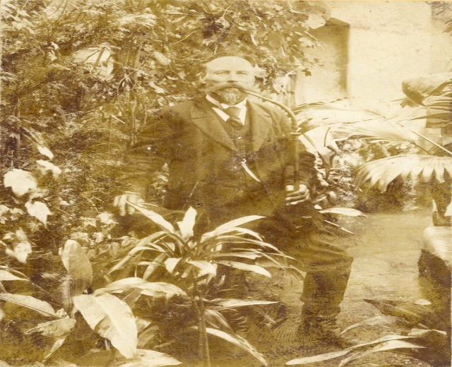 Altes Schwarz-Weiß-Foto eines Mannes in Anzug und Krawatte, der in einem Garten mit Pflanzen, Bäumen und einer Wand im Hintergrund steht.