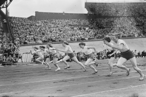 Eine Gruppe von Frauen, die auf einer Laufbahn in einem Stadion laufen, mit Zuschauern auf den Tribünen, dargestellt in Schwarz-Weiß.